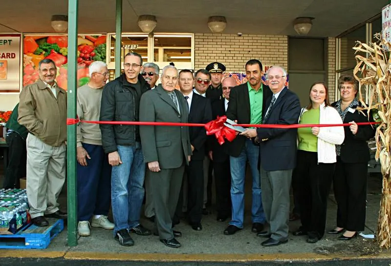Greenland Market A group of people stands in front of Greenland Market during a ribbon-cutting ceremony. A man holds scissors cutting the red ribbon, surrounded by others in formal attire and press from Square 4 Media capturing the moment. SQUARE 4 MEDIA